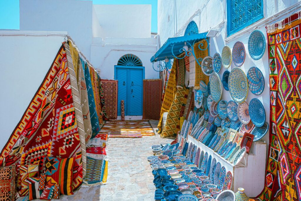 Um beco estreito em uma cidade do norte da África ou do Oriente Médio, ladeado por tapetes coloridos e cerâmicas decoradas penduradas para venda.