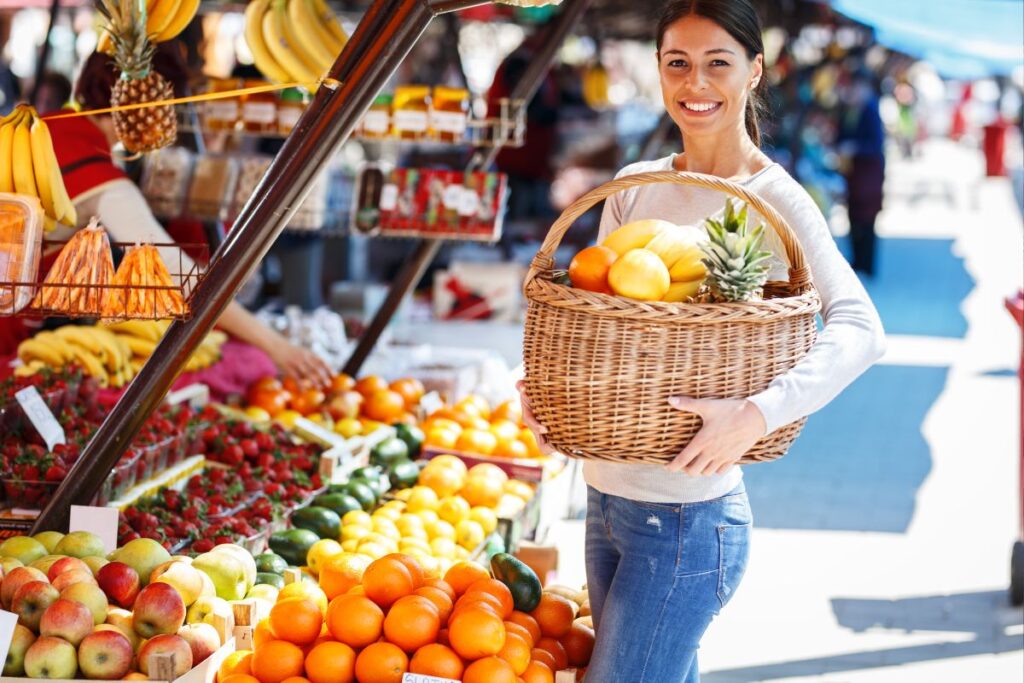 Uma mulher sorridente segurando uma cesta de frutas tropicais frescas em um mercado de rua ao ar livre com pilhas de frutas e vegetais.