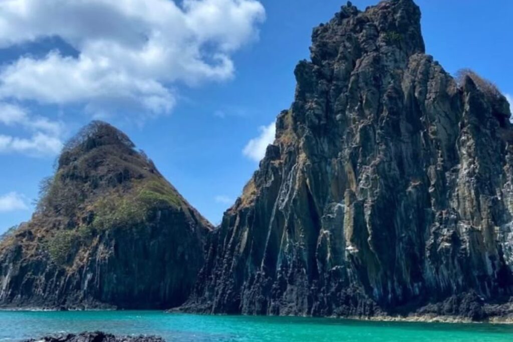 Vista panorâmica da Baía dos Porcos em Fernando de Noronha, com águas translúcidas em tons de verde e azul, areia dourada e os icônicos morros "Dois Irmãos" ao fundo sob um céu azul com nuvens.