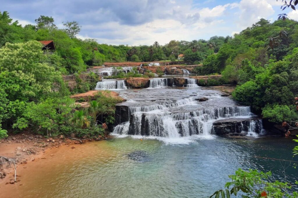 Cachoeiras em degraus com águas cristalinas formando piscinas naturais e uma pequena praia de areia, rodeadas por vegetação densa e exuberante, em Bonito, Mato Grosso do Sul.