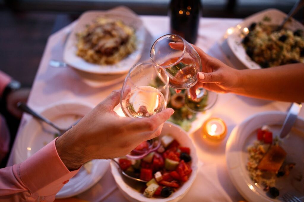 Brinde romântico de um casal com taças de vinho branco, cercado por pratos deliciosos e velas, em um jantar íntimo.
