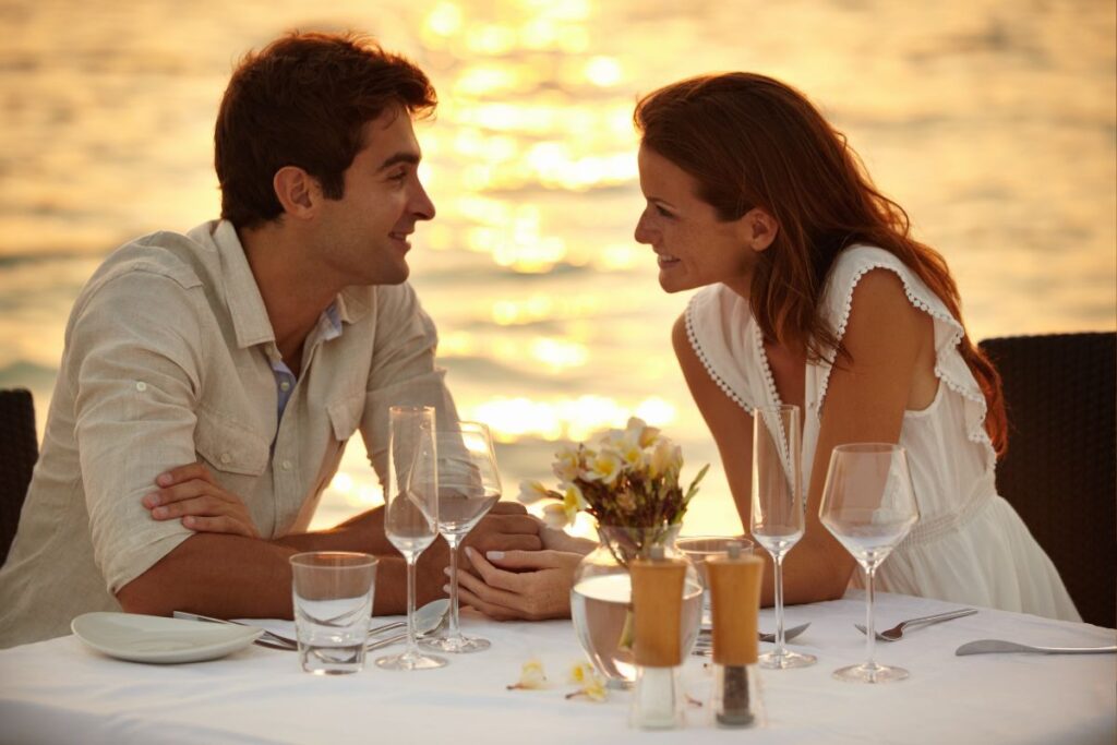 Casal sorridente em um jantar romântico ao pôr do sol, com o reflexo dourado na água ao fundo, em um cenário de viagem paradisíaco.