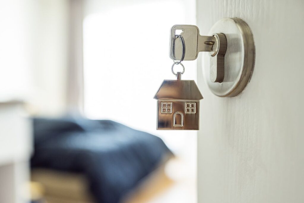 Chave de uma casa pendurada na fechadura de uma porta aberta, com um chaveiro em formato de casa.