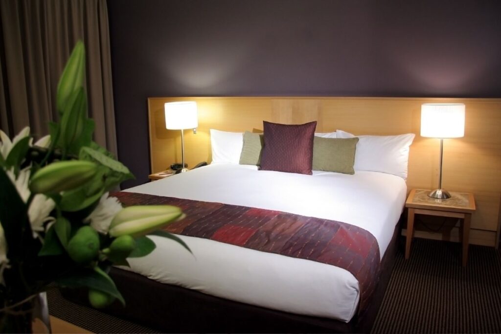 Quarto de hotel moderno com cama arrumada, travesseiros coloridos e luminárias nas mesas de cabeceira, oferecendo conforto em hospedagens.