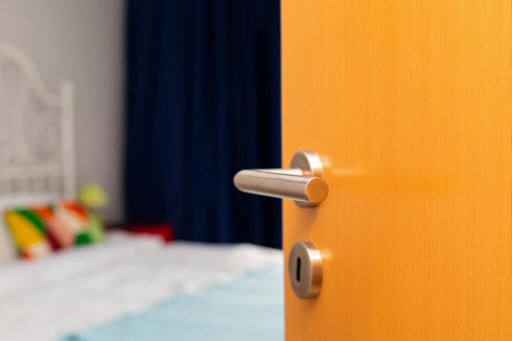 Porta de madeira aberta revelando um quarto de hotel claro com cama, indicando boas-vindas às hospedagens.