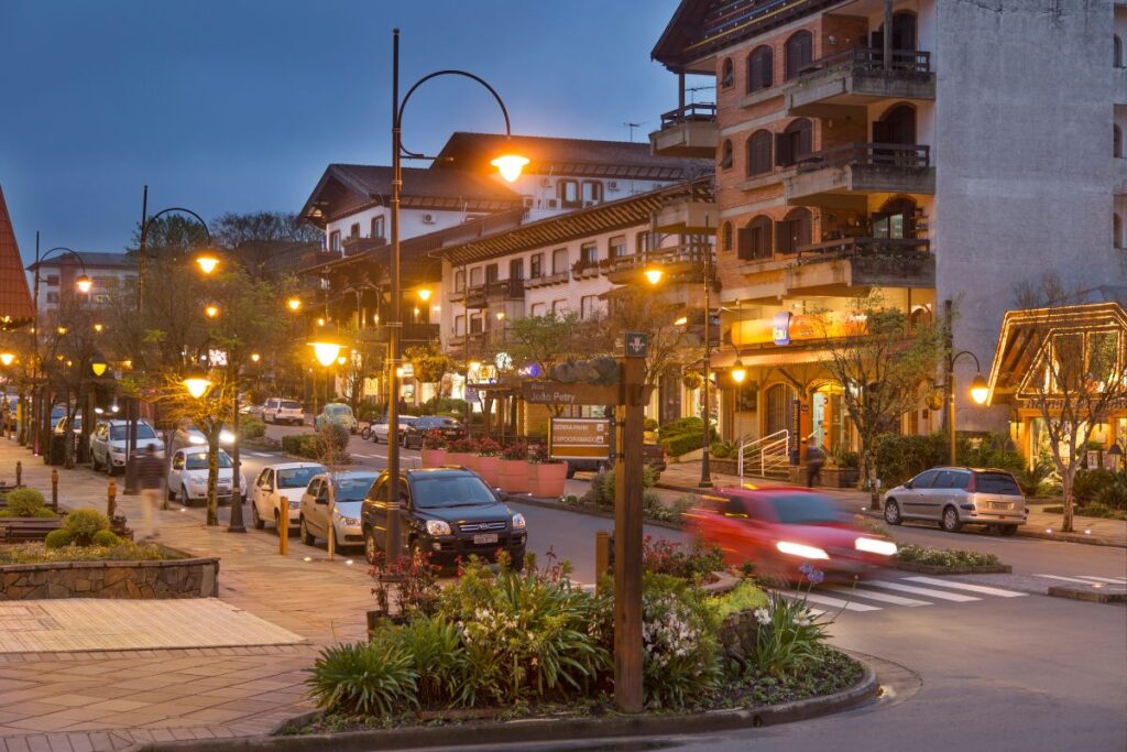 Uma vista noturna da rua principal de Gramado, com carros estacionados e pessoas caminhando sob a iluminação quente dos postes de luz e das fachadas dos edifícios. As luzes criam um ambiente acolhedor e convidativo para uma viagem romântica.