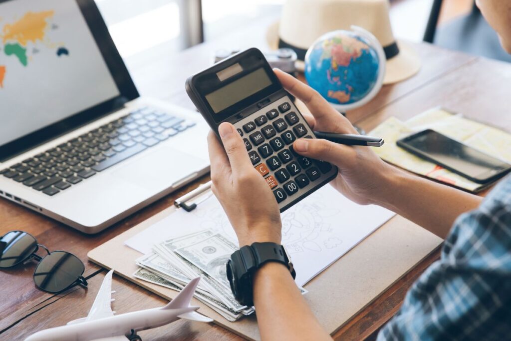 Pessoa calculando o orçamento de uma viagem com uma calculadora, notebook e mapa.