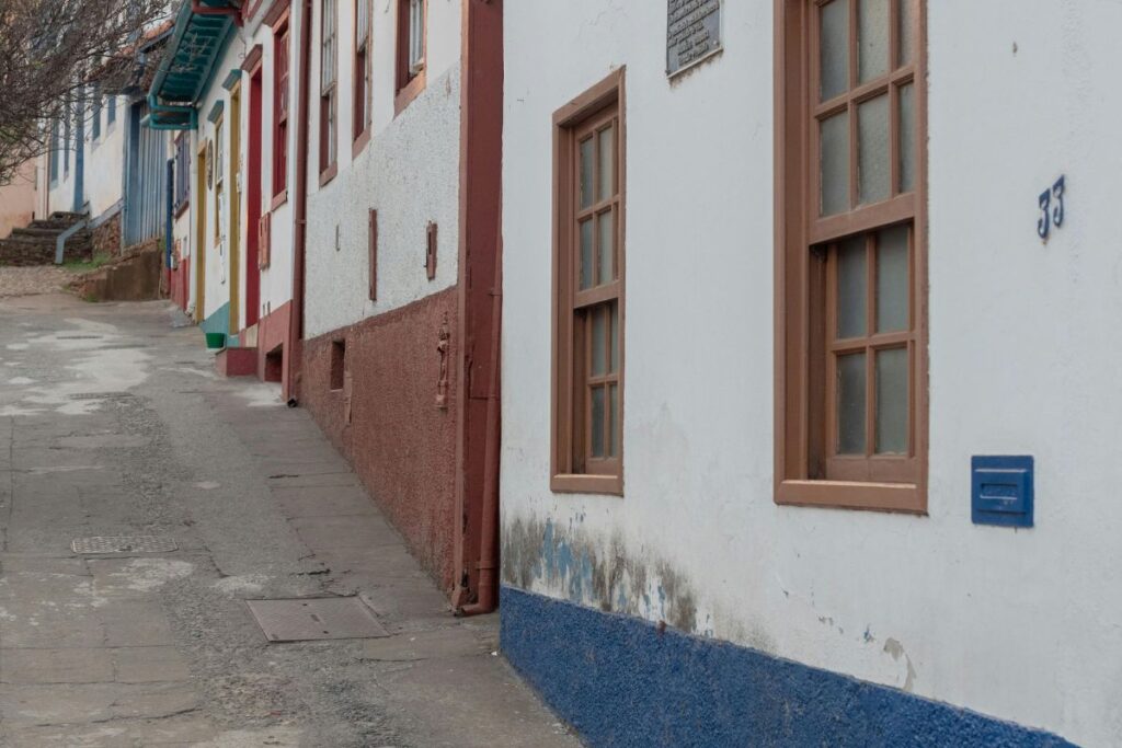 Rua estreita e inclinada em Sabará, ladeada por casas coloniais brancas com detalhes em marrom e azul, e janelas de madeira. A paisagem urbana histórica e bem preservada é visível.