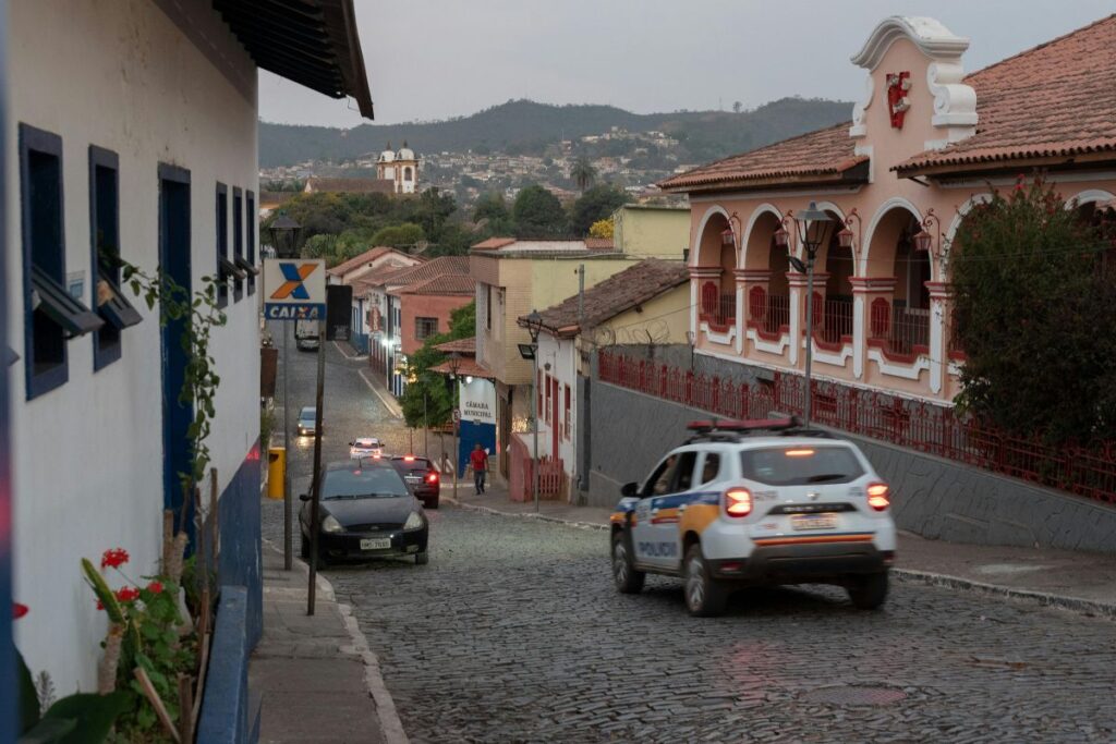 Rua de paralelepípedos em Sabará com uma viatura policial em primeiro plano e edifícios coloniais de arquitetura barroca nas laterais. Ao fundo, a Igreja do Carmo e a serra complementam a paisagem.