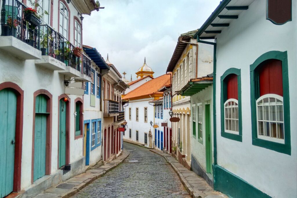 Vista de uma rua de paralelepípedos em Sabará, com casarões coloniais de diferentes cores, janelas e portas bem preservadas. A rua se curva em direção ao fundo, onde é possível ver o telhado de uma igreja.