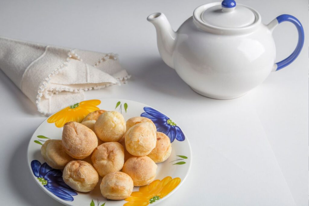 Um bule de chá branco e azul ao lado de um prato com pães de queijo, sobre uma superfície branca.