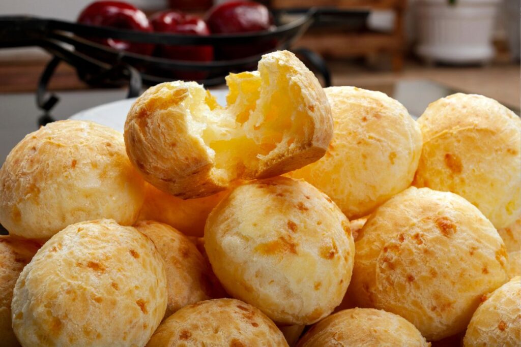 Pães de queijo dourados e crocantes empilhados, com um deles aberto mostrando o interior macio e elástico, o verdadeiro pão de queijo.