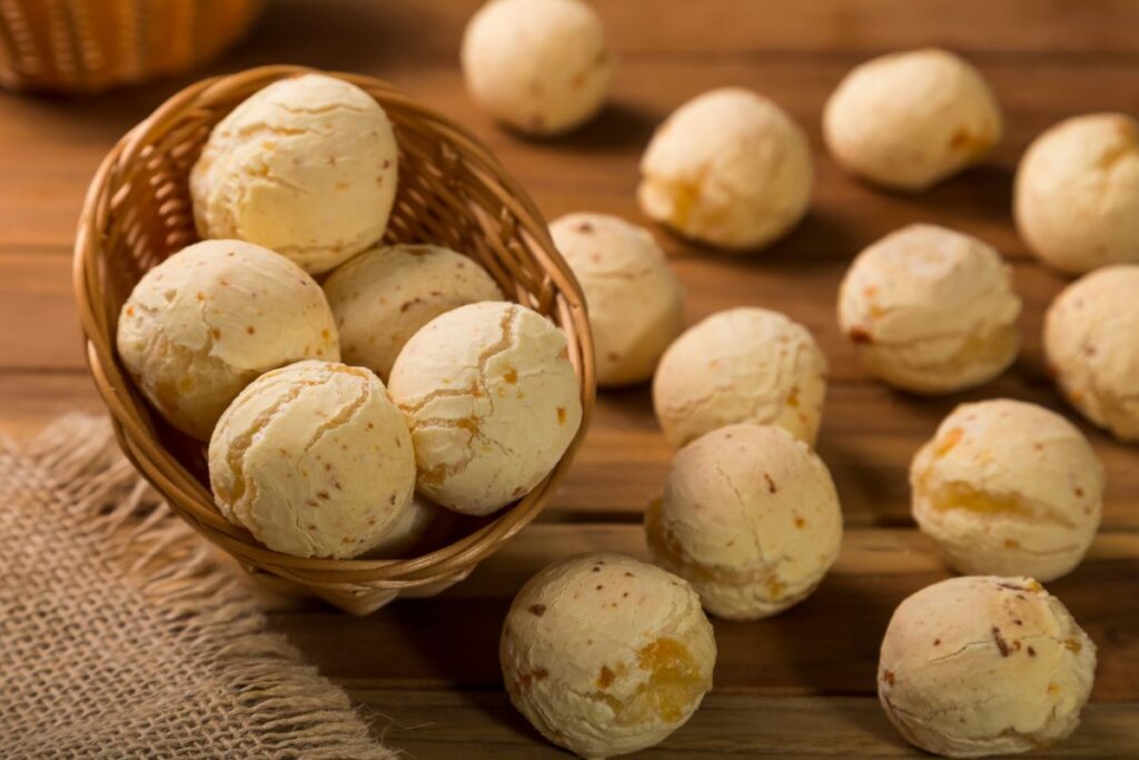 Pães de queijo macios e dourados, alguns dentro de uma cesta de vime e outros espalhados sobre uma mesa de madeira. A cena evoca a simplicidade e o acolhimento de Minas Gerais.