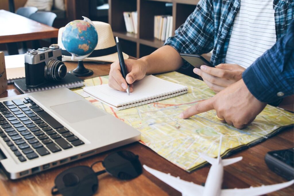 Duas pessoas trabalhando juntas em um roteiro de viagem, com laptop, mapa e caderno.
