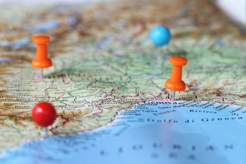 Roteiro de viagem sendo planejado em um mapa antigo, com alfinetes de diferentes cores marcando cidades como Gênova e Riviera.