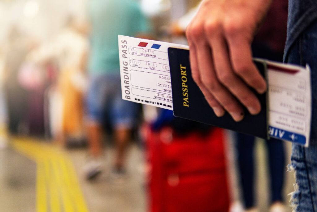 Mão segurando passaporte e cartão de embarque em um aeroporto movimentado, o que representa o início da viagem após o planejamento do roteiro.