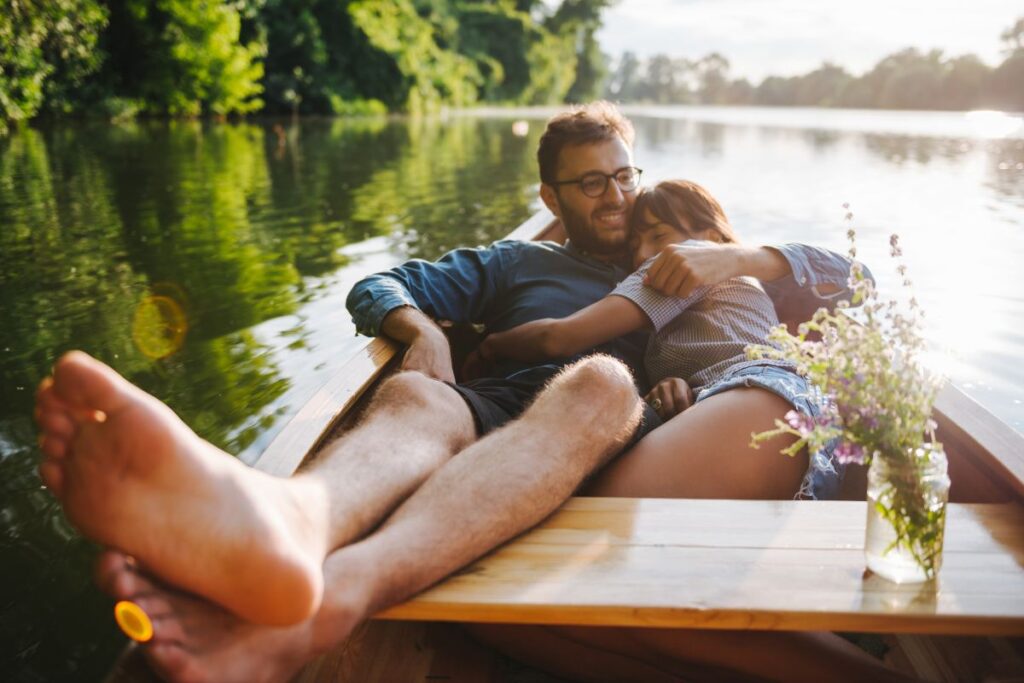 Casal abraçado em um passeio romântico de barco a dois em um lago tranquilo ao pôr do sol, cercado por árvores verdes.