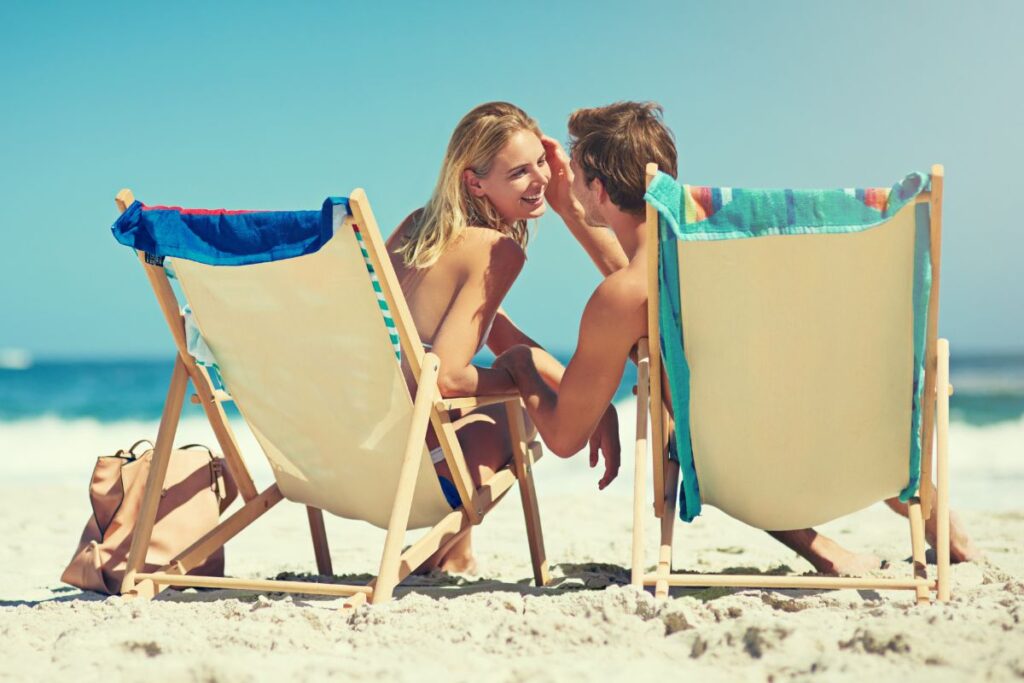 Casal em um passeio romântico a dois na praia, aproveitando um momento de cumplicidade em cadeiras de praia sob o sol e céu azul.

