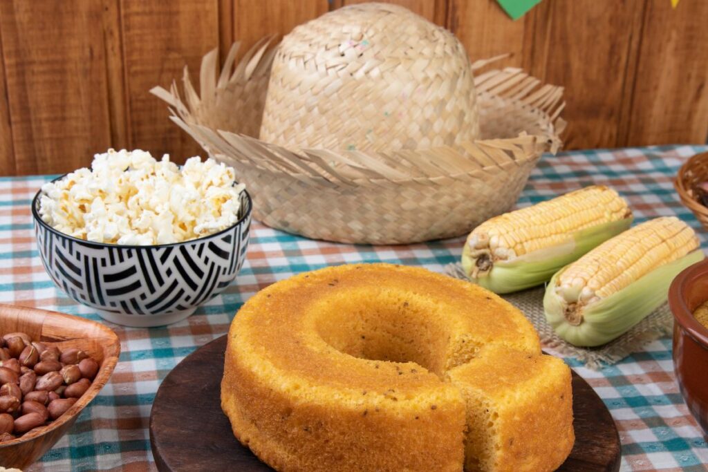Mesa com comidas típicas de festa junina: bolo de fubá, pipoca, milho, amendoim e chapéu de palha sobre toalha xadrez, tradições das melhores festas de São João do Brasil