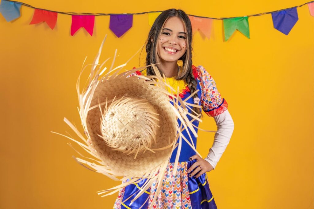 Jovem sorridente com vestido caipira colorido segurando chapéu de palha em cenário de festa junina com bandeirinhas, representando as tradições do São João brasileiro