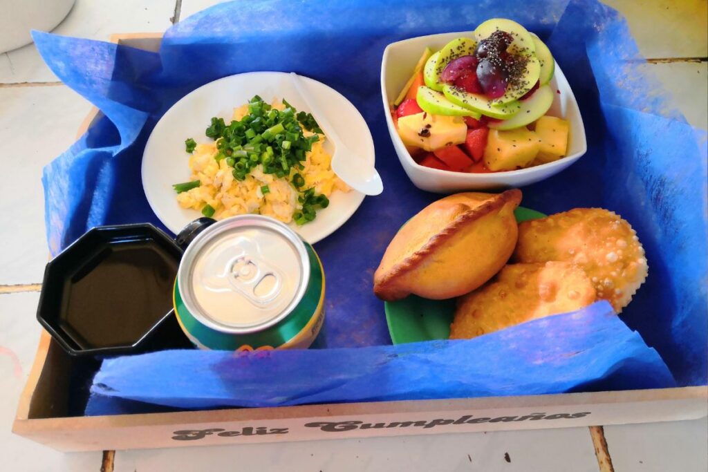 Uma bandeja de café da manhã de aniversário com ovos mexidos, salada de frutas, empanadas e uma bebida.