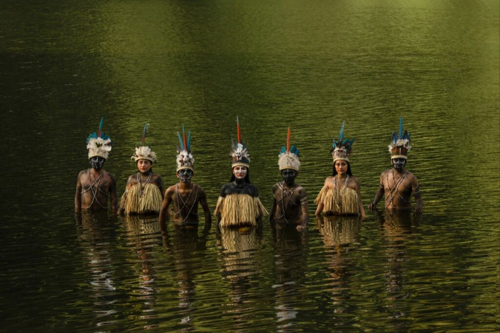 Grupo indígena em ritual tradicional nas águas, usando pinturas corporais e adornos que simbolizam ancestralidade e conexão com a natureza.