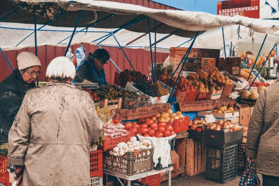 Pessoas comprando frutas e vegetais frescos em um mercado de rua coberto, com caixas de produtos agrícolas variados.