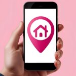 Mão segurando um smartphone com um ícone de pino de localização e uma casa dentro, em fundo rosa.
