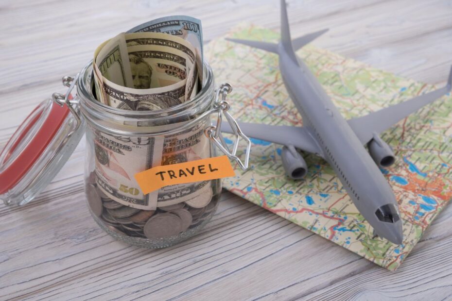 Jarrinho de vidro com moedas e notas de 100 dólares e um rótulo escrito 'travel' ao lado de um avião de brinquedo e um mapa. A imagem representa o planejamento de viagens econômicas.