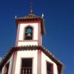 Close-up do campanário da Igreja de Nossa Senhora do Ó em Sabará, com suas paredes brancas e detalhes em bordô. Um sino azul e um crucifixo no topo se destacam contra o céu azul sem nuvens.