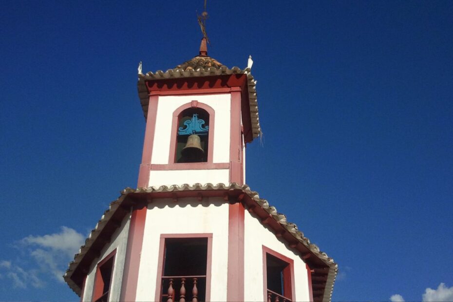 Close-up do campanário da Igreja de Nossa Senhora do Ó em Sabará, com suas paredes brancas e detalhes em bordô. Um sino azul e um crucifixo no topo se destacam contra o céu azul sem nuvens.
