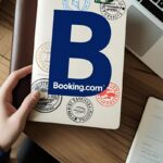 Mão segurando caderno com logo Booking.com e selos, laptop e café na mesa. Planejamento de viagens online Booking.com.