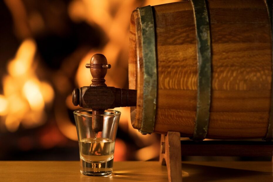 Barril de cachaça de madeira com torneira servindo dose em copo shot, ambiente rústico com fogo ao fundo.