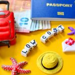 Vários itens de viagem, incluindo um passaporte, uma mala pequena, uma bússola e notas de euro, com blocos de letras formando a frase "LOW COST".