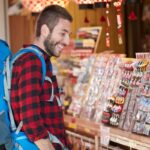 Turista com mochila observando e sorrindo para a seleção de artesanato local em uma loja, buscando uma conexão cultural através de produtos artesanais.