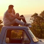 Casal aproveitando um passeio a dois ao pôr do sol sentado no teto de uma caminhonete com vista para o mar