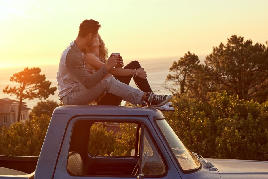 Casal aproveitando um passeio a dois ao pôr do sol sentado no teto de uma caminhonete com vista para o mar